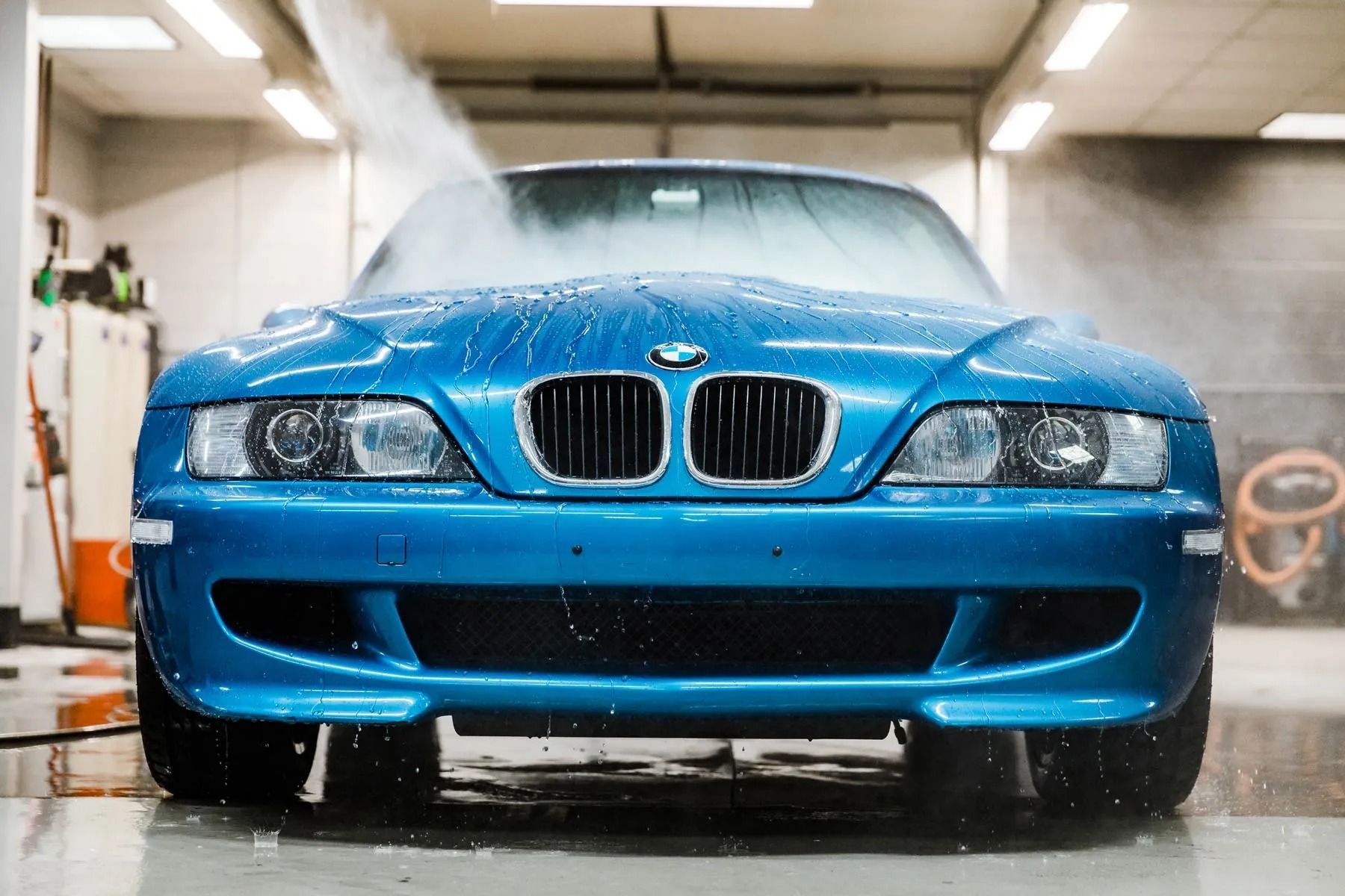 Blue BMW sedan being washed in a car wash, front view with water spraying.