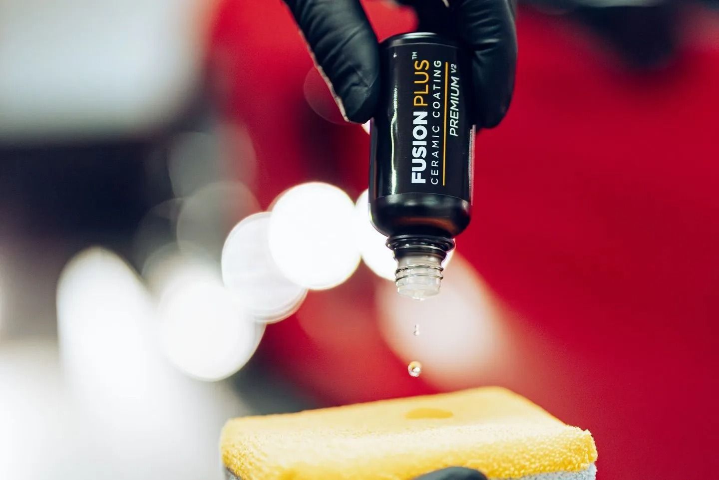 Gloved hand holding a black bottle dripping liquid onto a yellow sponge, with blurred red background