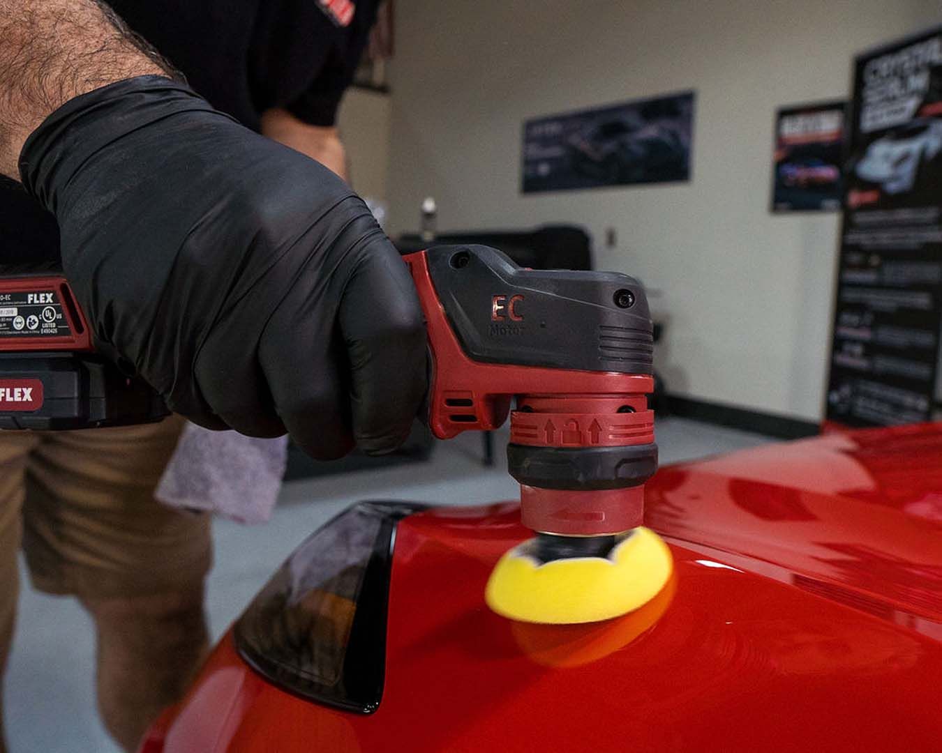 Gloved hand polishing a red car with a buffer and yellow pad