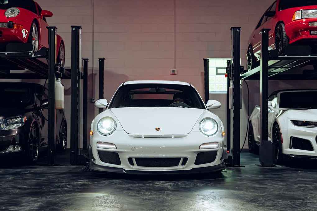 White sports car in a garage, flanked by red and white cars on lifts.