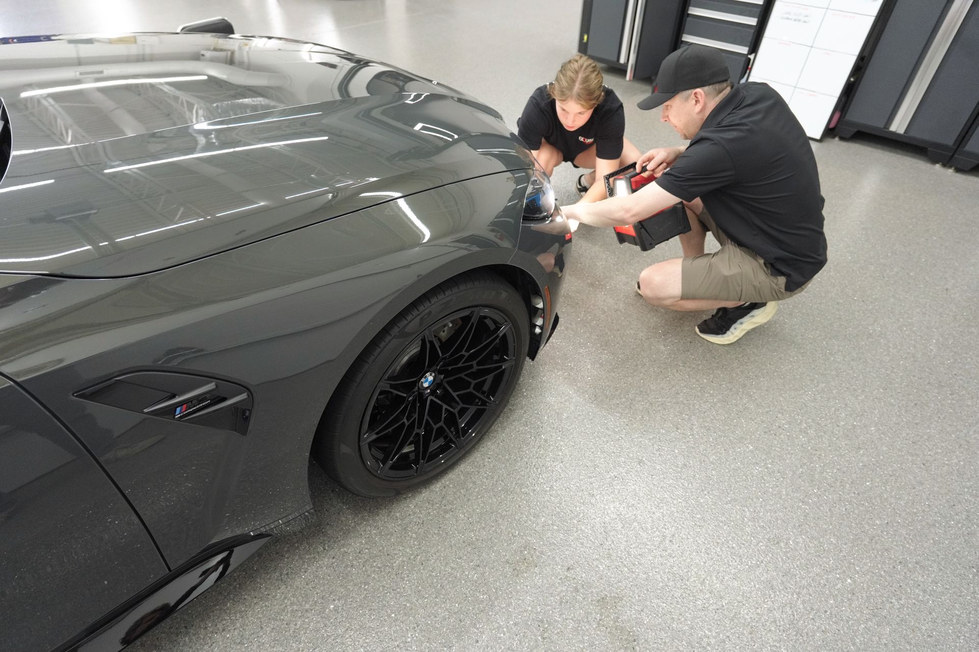 Two people crouch beside a gray sports car, inspecting the front wheel in a garage.