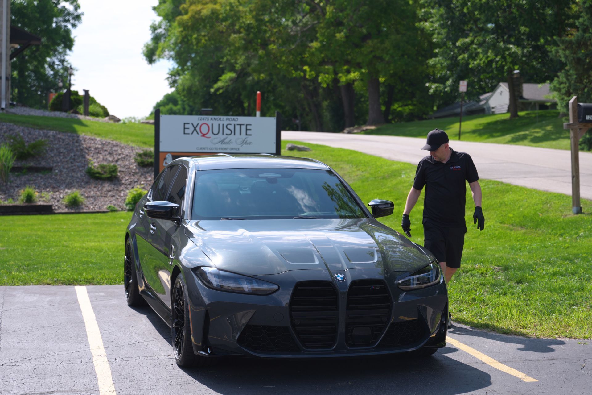 Black sports sedan parked by a driveway sign, with a man standing beside it on a sunny day