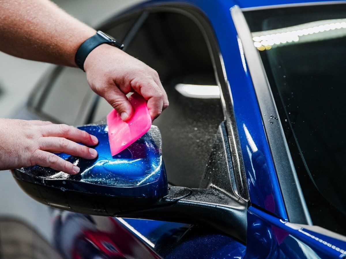 Hands cleaning a blue car side mirror with a pink sponge at a car wash
