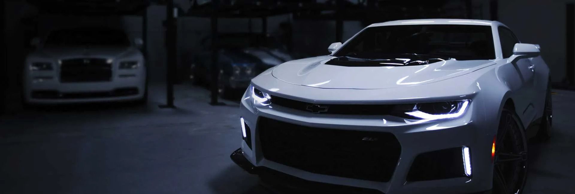 White sports cars parked in a dim garage, front car brightly lit with headlights on