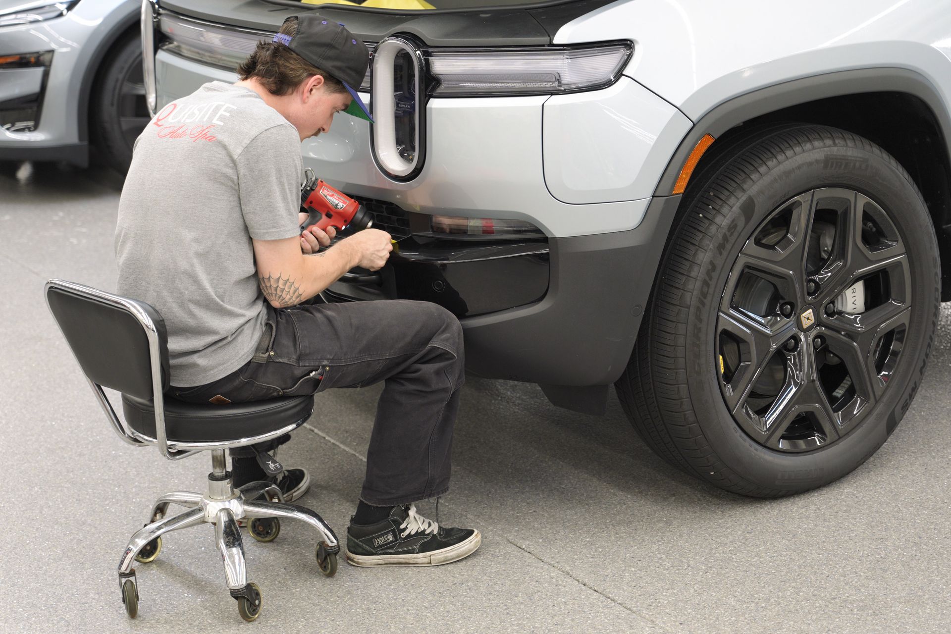 Person using a red power tool to repair the front bumper of a silver SUV while seated on a rolling chair.