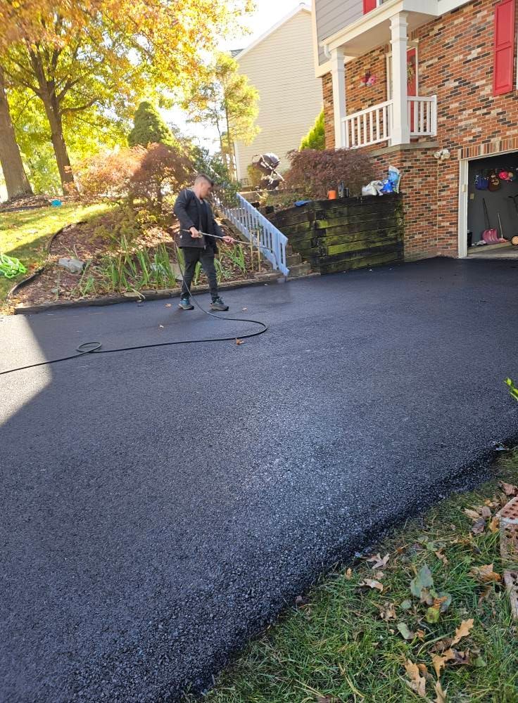 A person applying sealant to a newly paved asphalt driveway in front of a brick house.