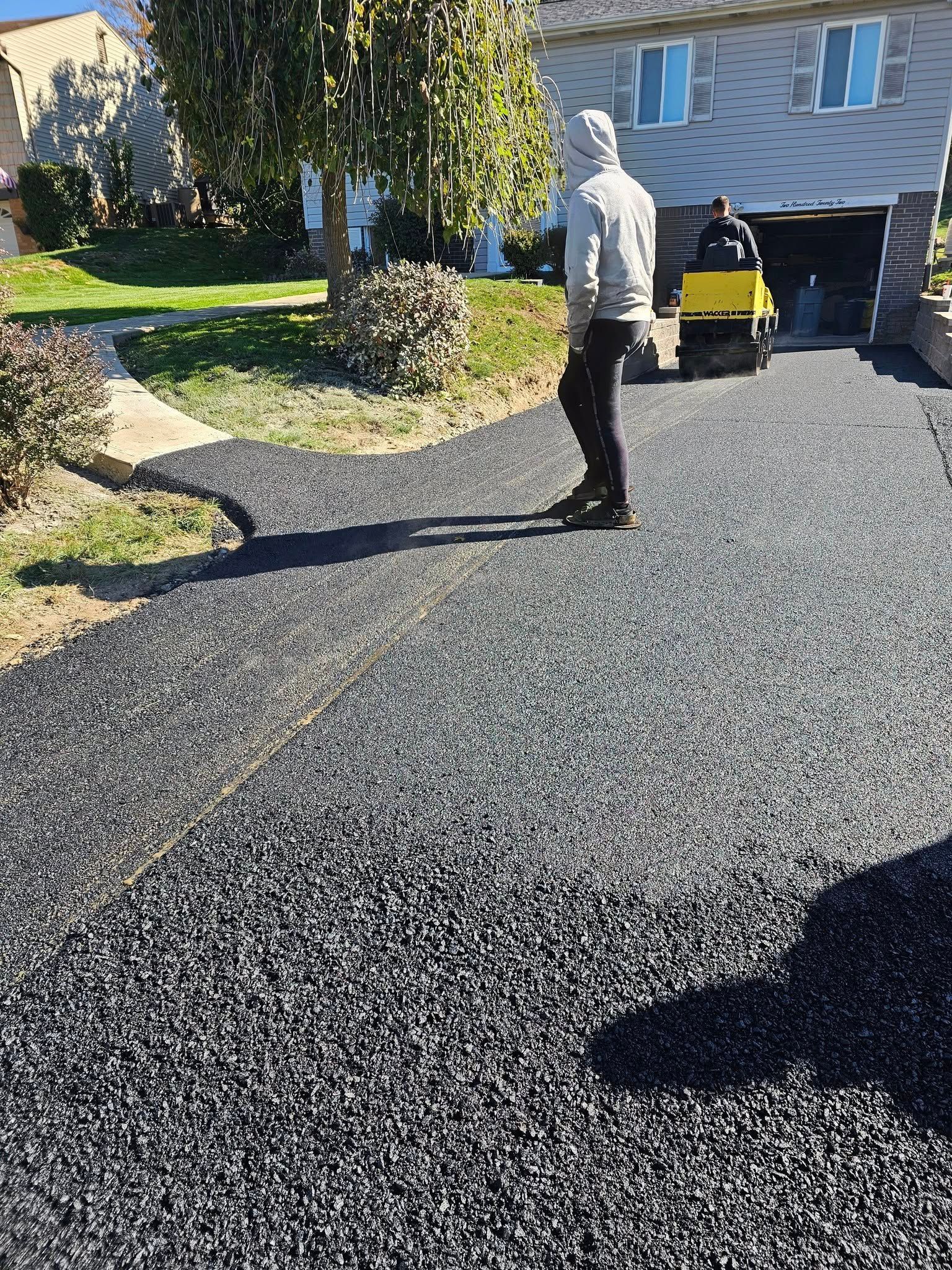 A person in a gray hoodie walks on a newly paved asphalt driveway in front of a house, near a small construction roller.