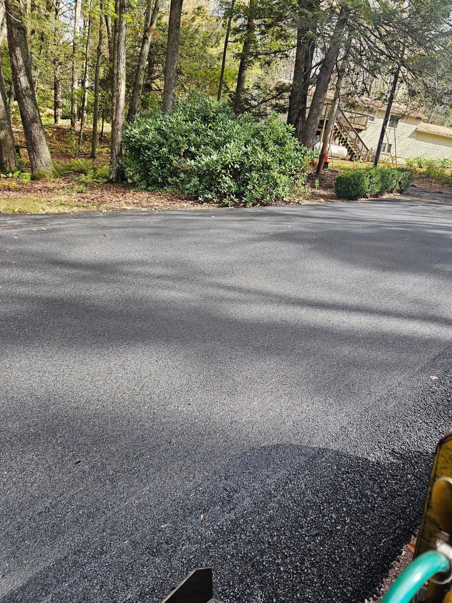 A view of a freshly laid asphalt driveway with a gravel texture, surrounded by trees and a green hedge.