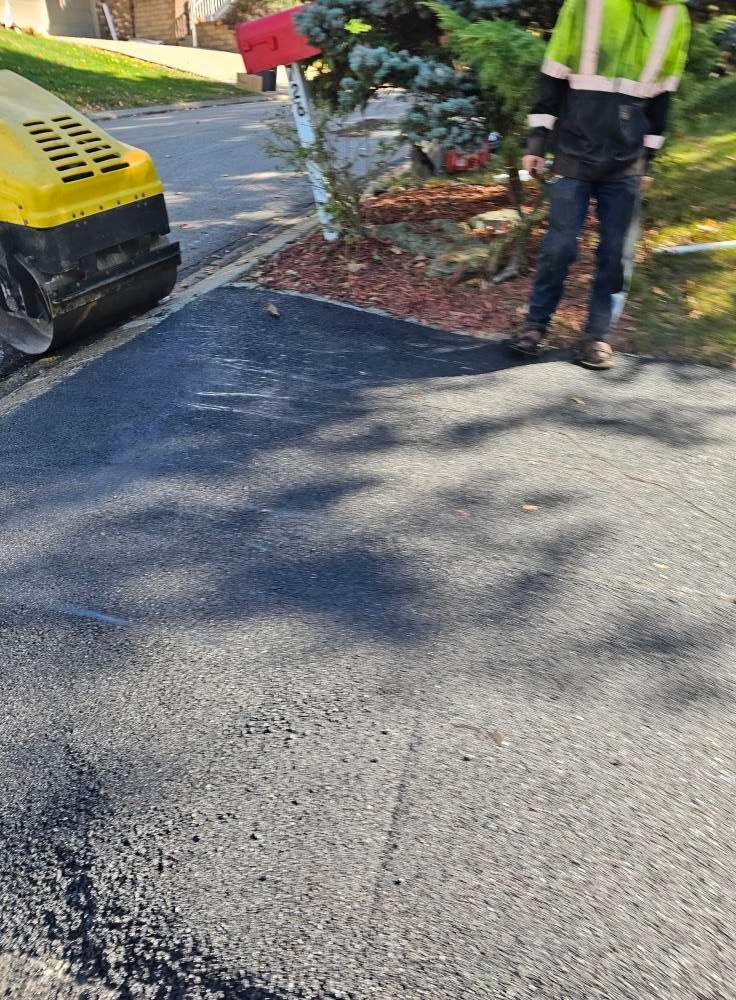 A construction worker in a high-visibility vest stands next to a road roller on a freshly paved asphalt driveway.
