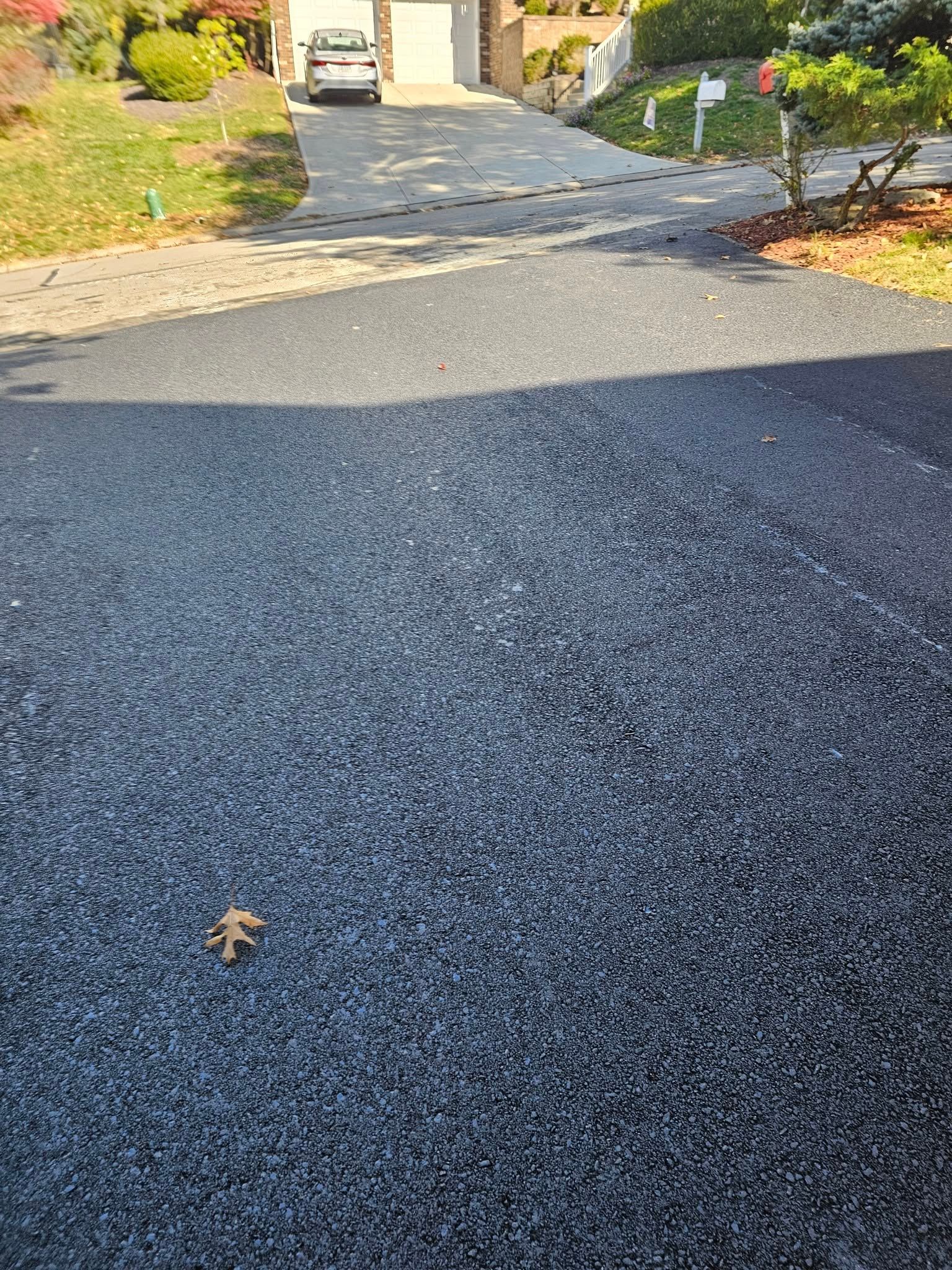 A view of a freshly paved black asphalt driveway leading toward a house garage with a parked car in the distance.