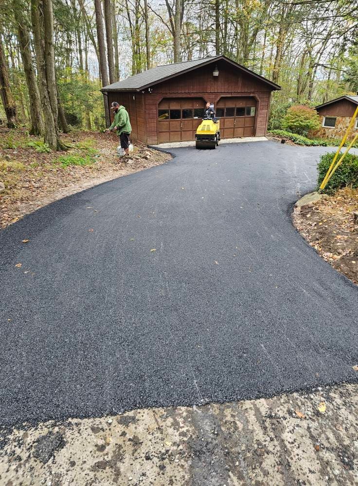 A person uses a yellow asphalt roller on a newly paved driveway in front of a wooden garage surrounded by trees.