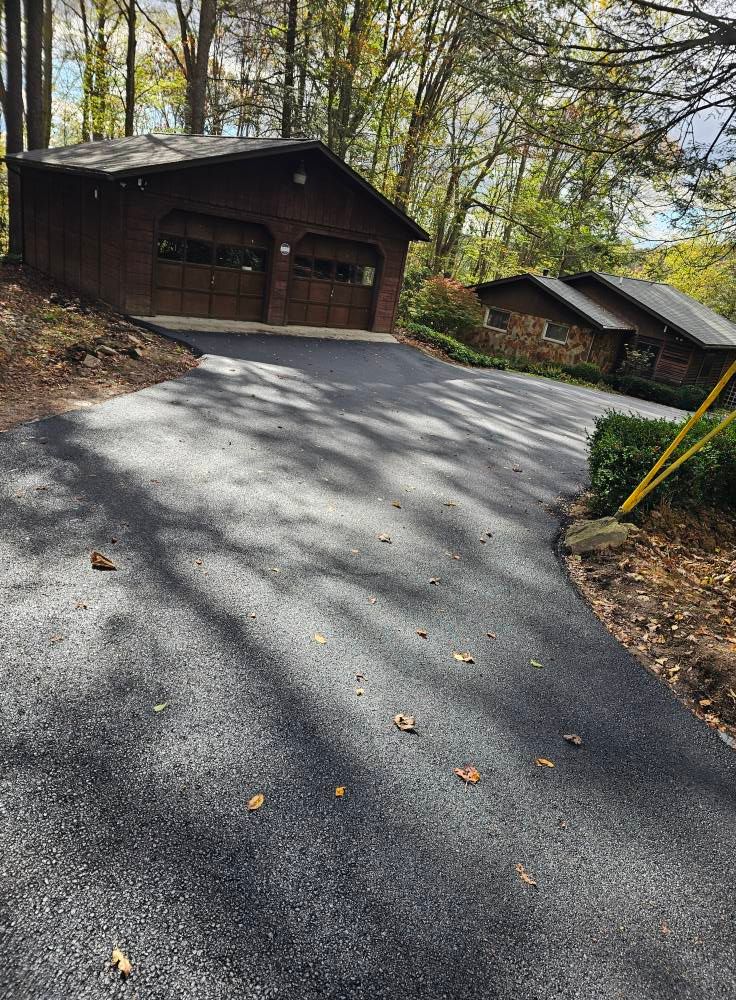 A freshly paved asphalt driveway leading to a brown two-car garage and a stone house surrounded by trees.