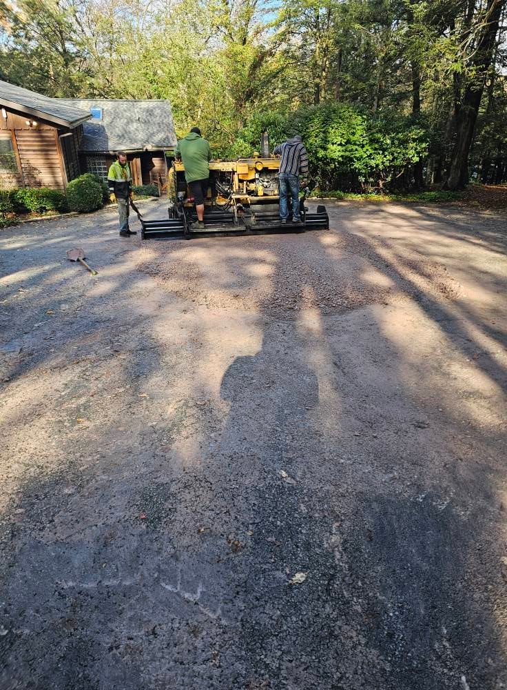 Three workers operate a yellow tractor to grade a gravel driveway in front of a house on a sunny day.