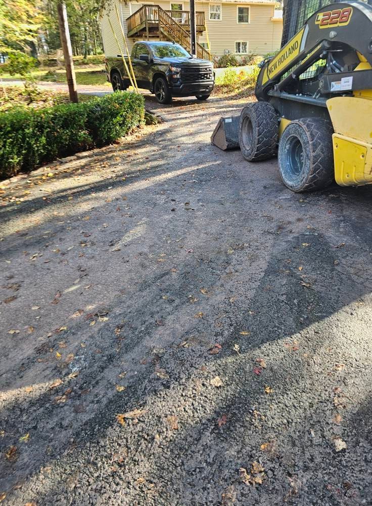 A yellow skid-steer loader parked on a gravel driveway near a black pickup truck and a house.