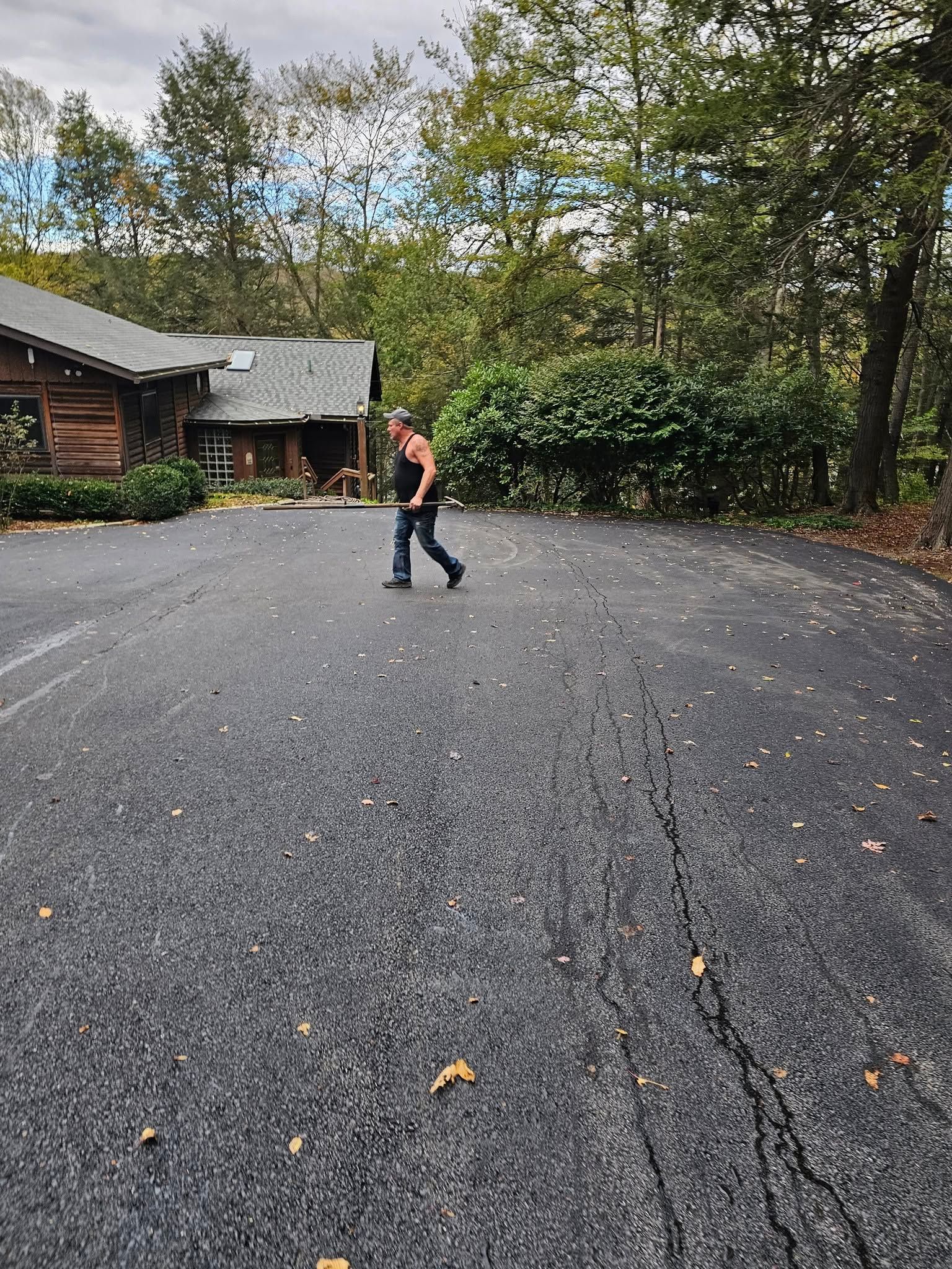 A person walks across a large, cracked asphalt driveway toward a log-style building surrounded by trees.