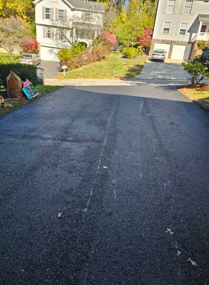 A freshly paved asphalt driveway leading toward two houses on a sunny, tree-lined suburban street.