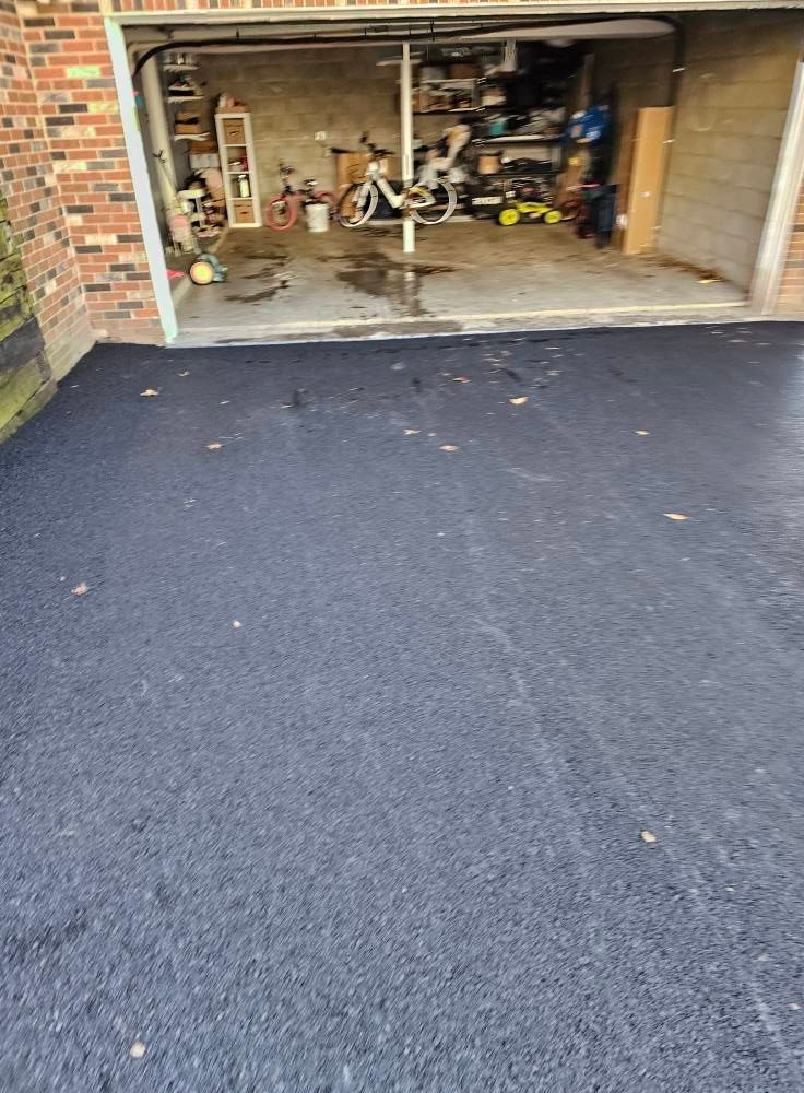 A freshly paved black asphalt driveway leads to a garage containing bicycles, storage shelves, and boxes.