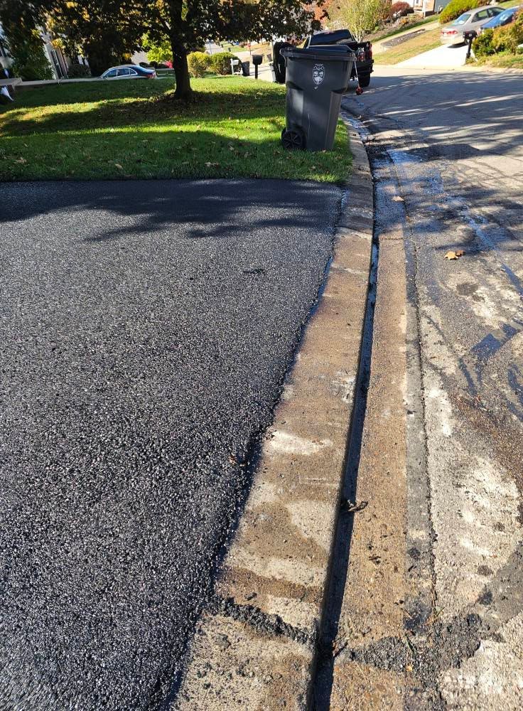 A fresh asphalt patch meets an existing concrete curb alongside a neighborhood street.