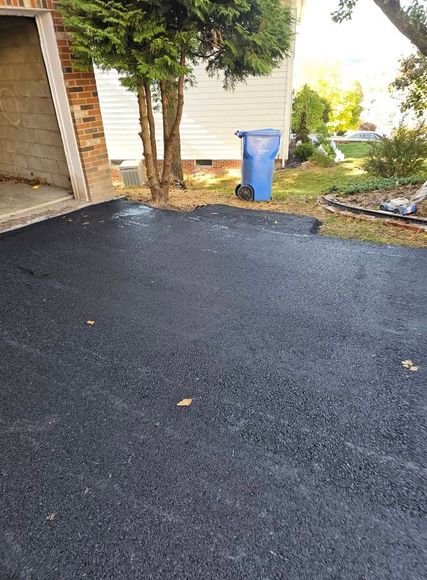 A freshly paved black asphalt driveway leading to a garage next to a tree and a blue trash bin.