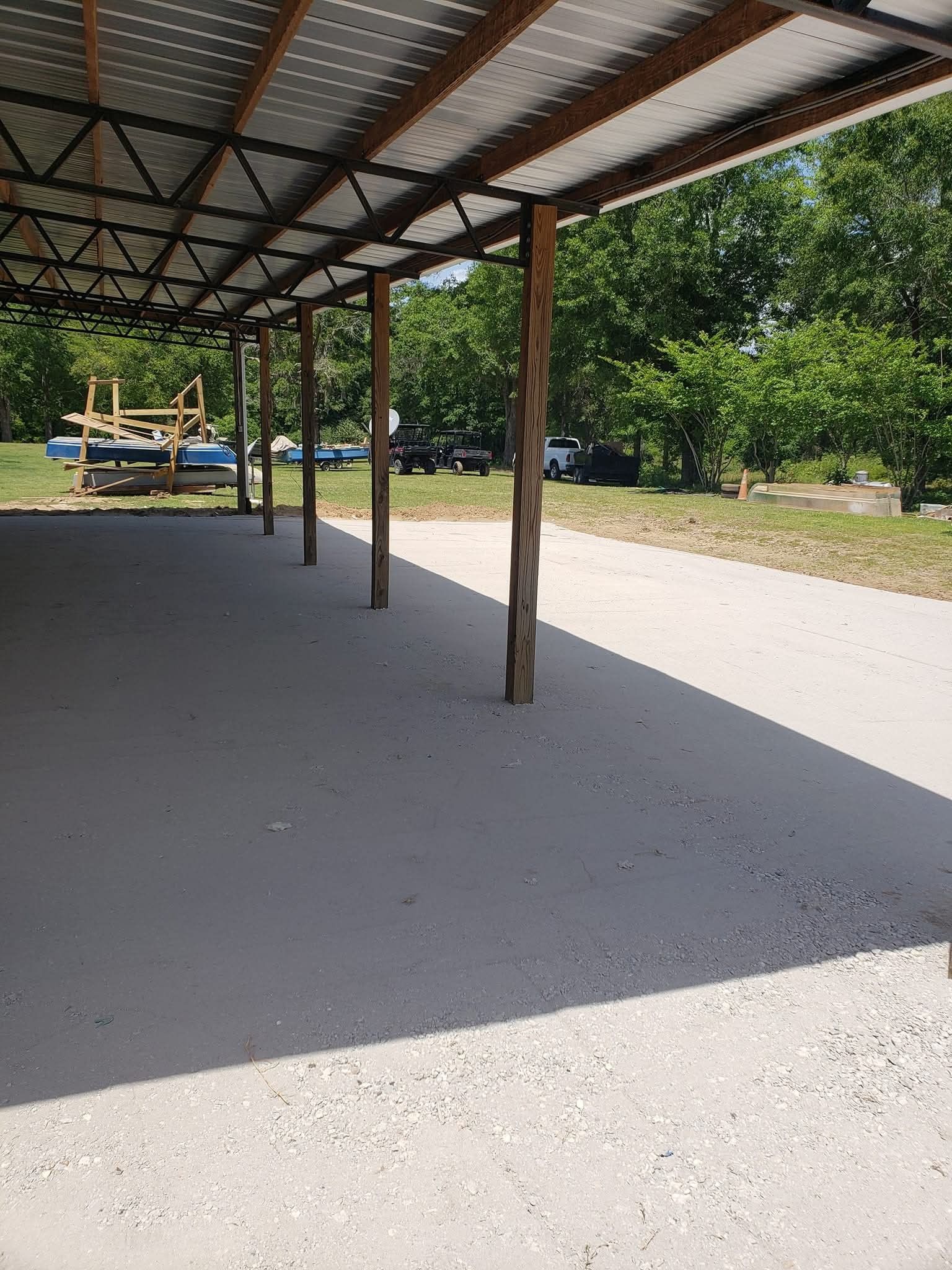 A view under a metal-roofed open shed with wooden support posts, overlooking a gravel area and distant green trees.