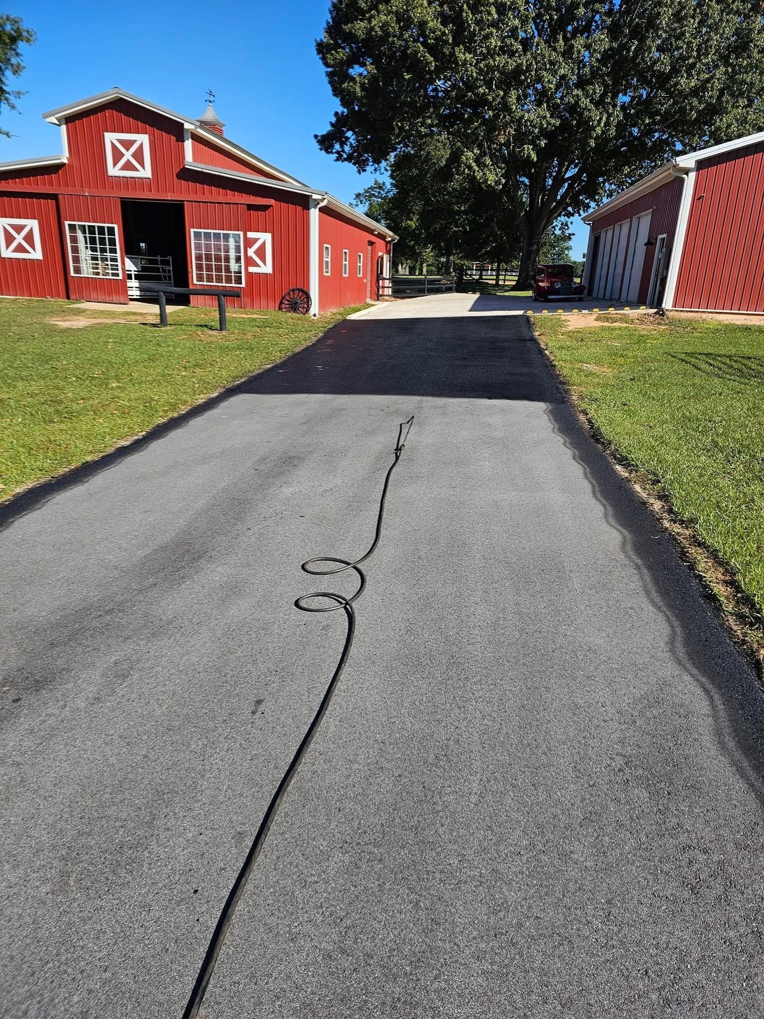 A paved driveway leads toward two red barns on a sunny day with green grass and a large tree in the background.