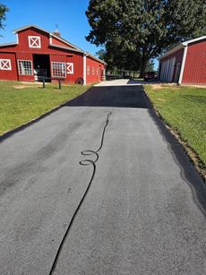 A paved driveway leads toward two red barns on a sunny day with green grass and a large tree in the background.