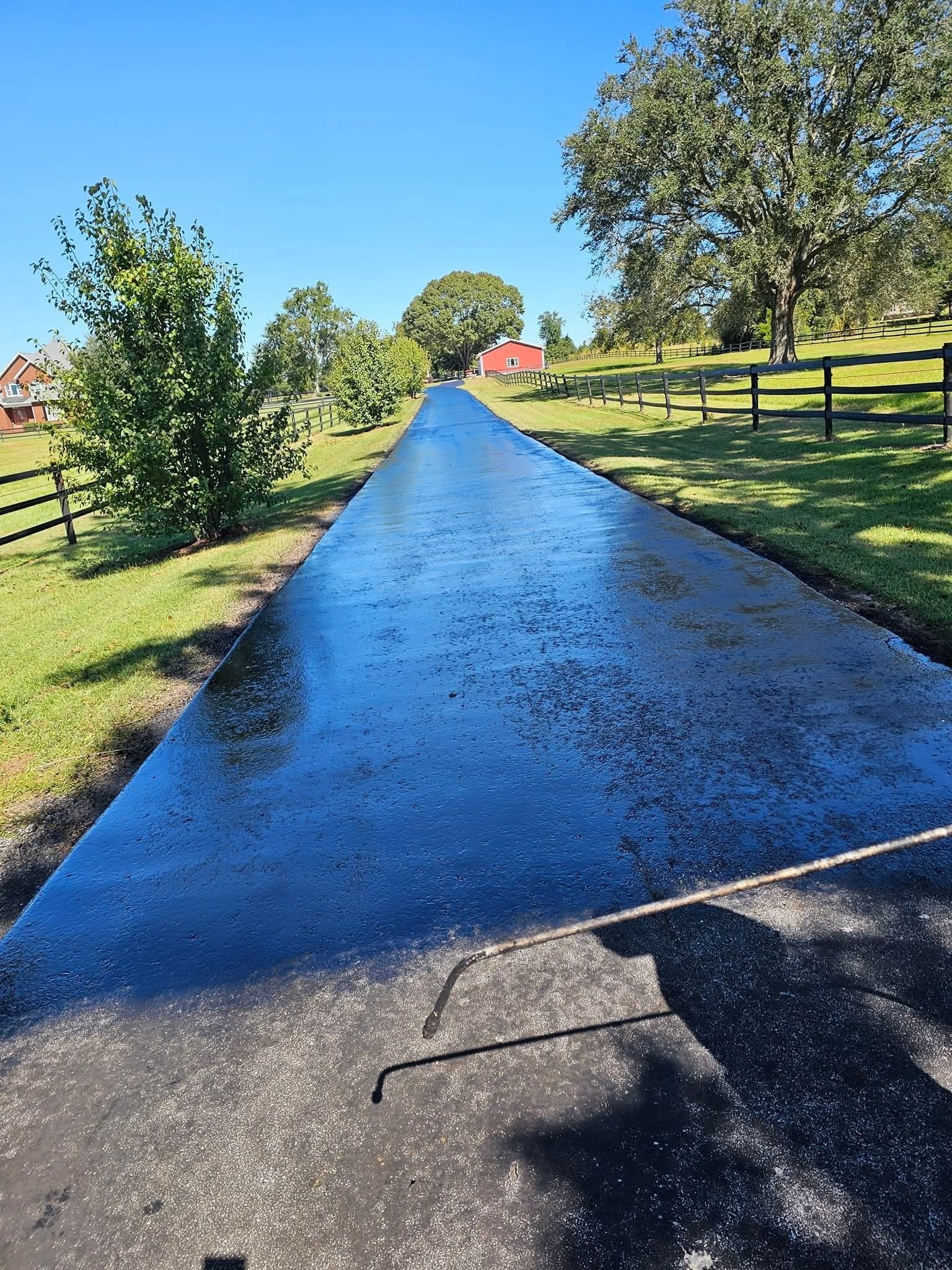 A freshly sealed, dark black asphalt driveway stretches between green grassy lawns and trees under a clear blue sky.