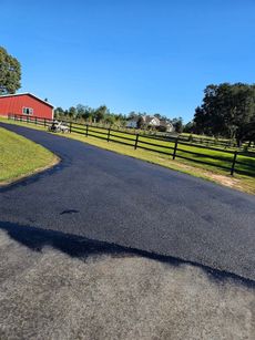 Freshly paved asphalt driveway leading to a red barn under a clear blue sky on a sunny day.