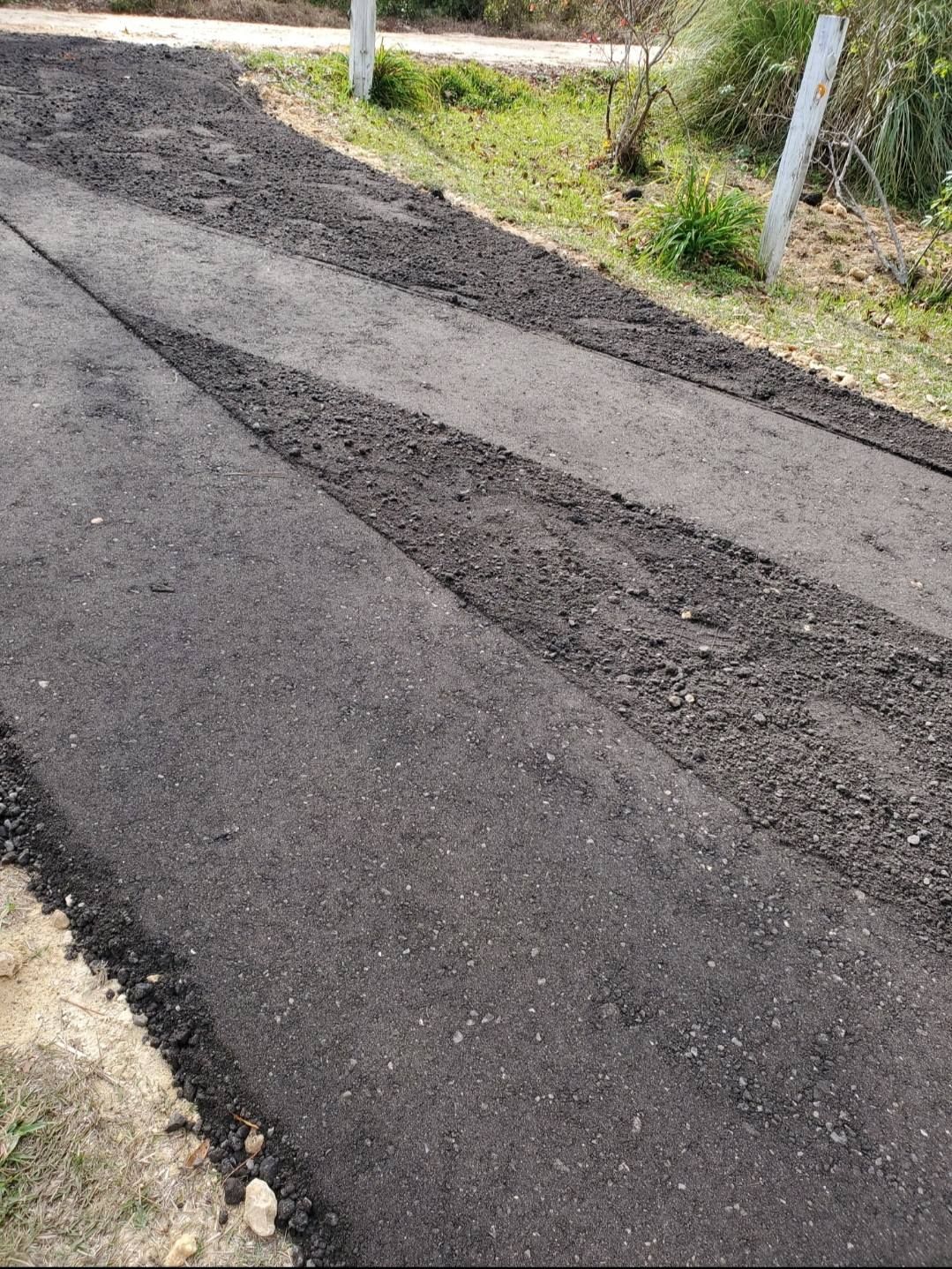 A path of dark, freshly laid asphalt with a strip of rougher gravel cutting across it, situated next to a grassy area.