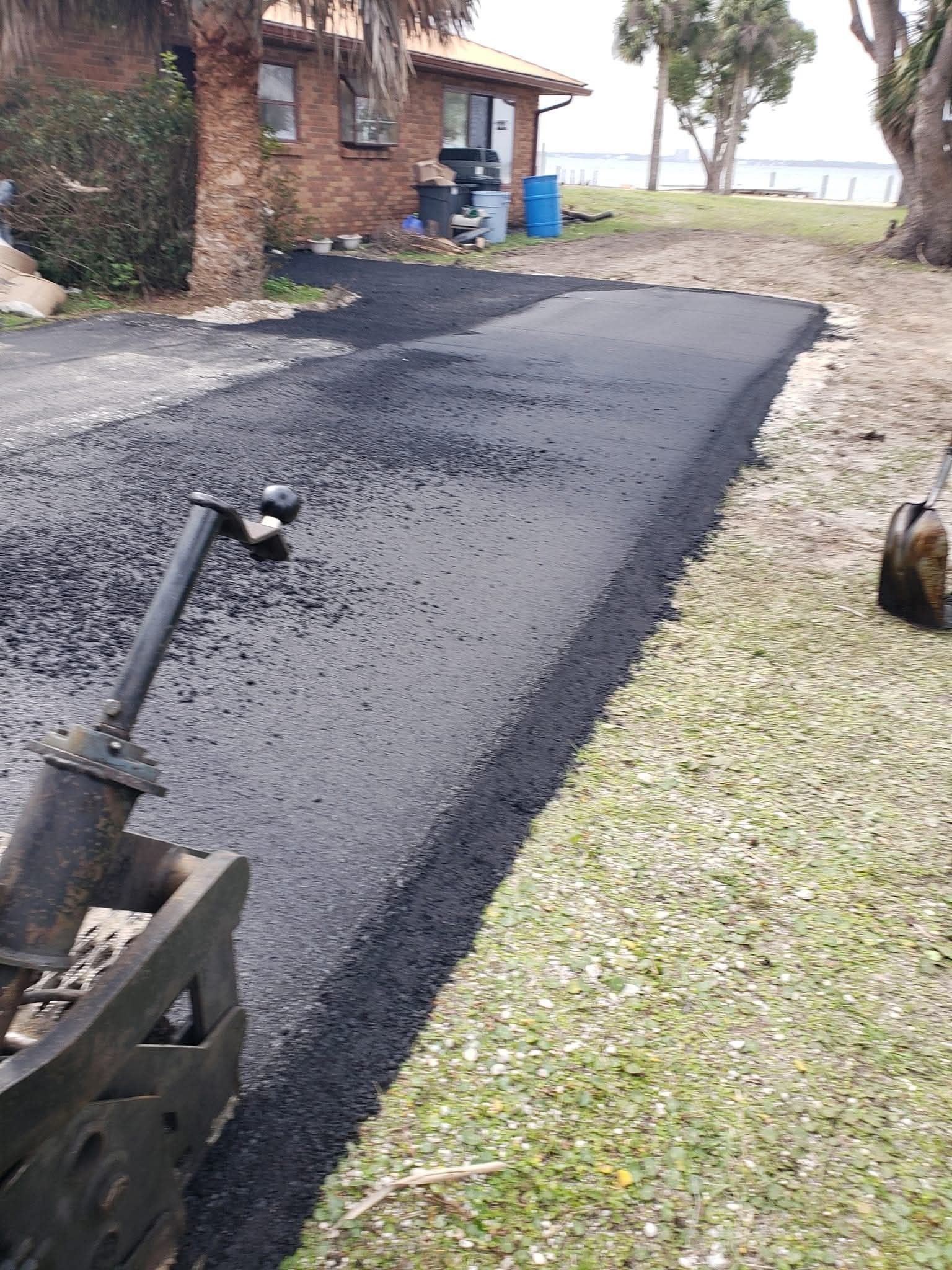 A freshly laid asphalt driveway leading to a brick house on a sunny day, with a heavy roller visible in the foreground.