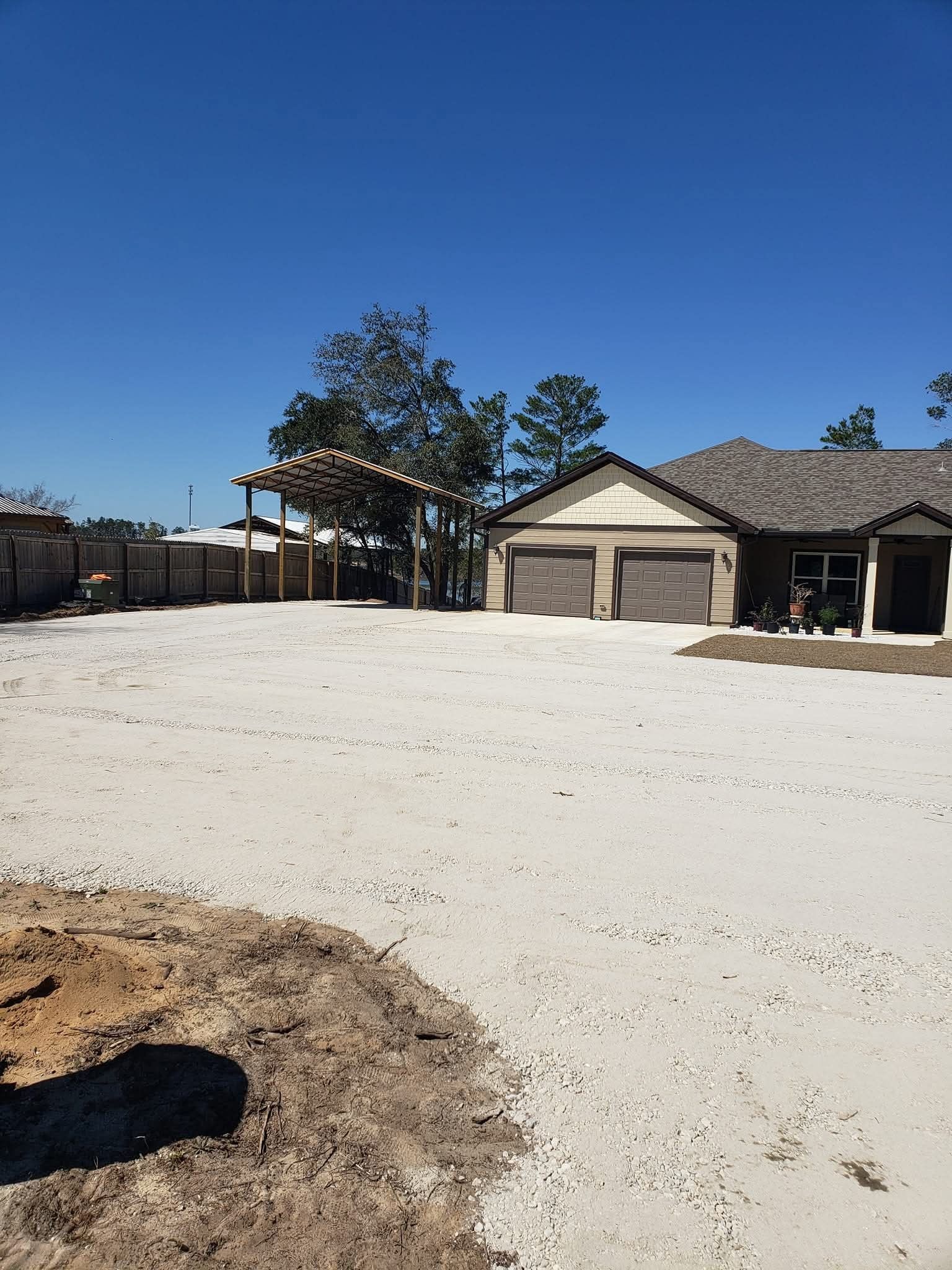 A sunny, wide-angle view of a house with a large, light-colored gravel driveway, a wooden pergola, and a clear blue sky.