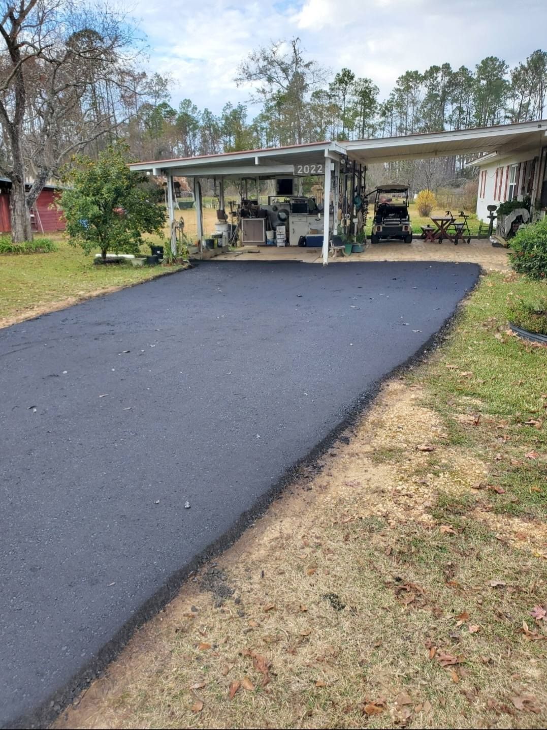 A newly paved, dark asphalt driveway leads to a covered carport structure in a residential yard.