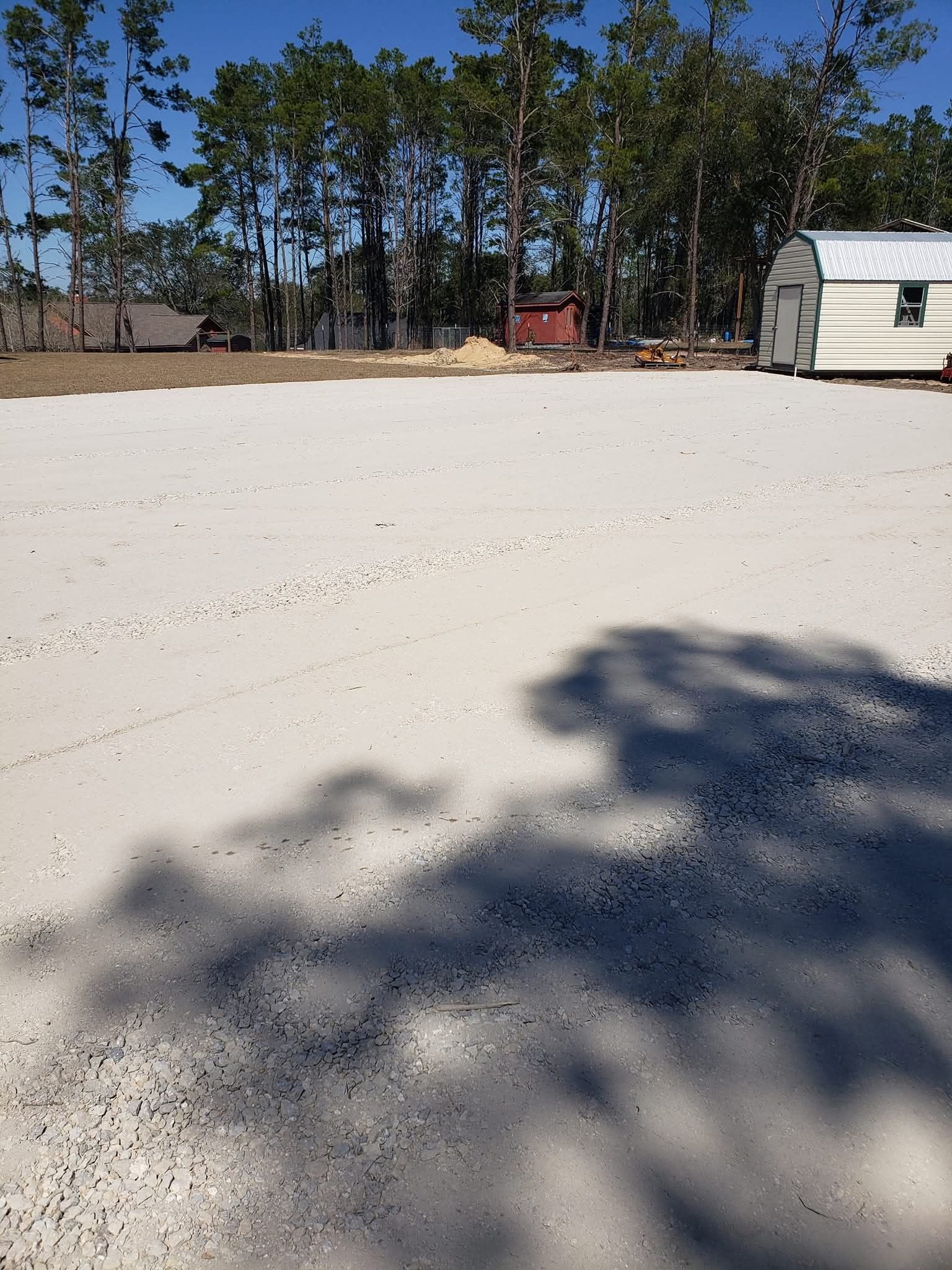 A large gravel lot under a clear blue sky, bordered by trees and a small storage building.