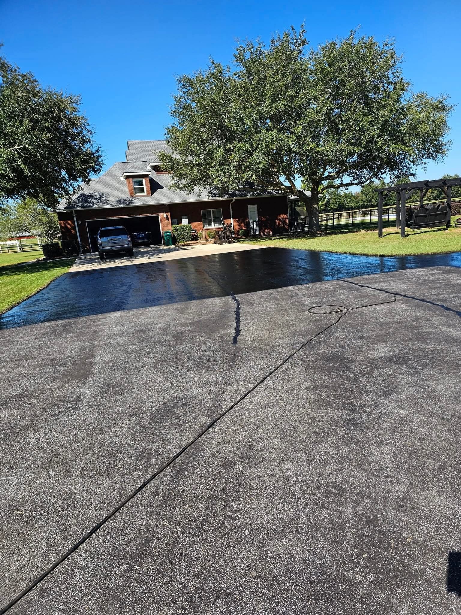 A paved driveway leads to a brown house under a clear blue sky, with large trees on both sides of the lawn.