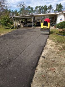 A person operates a yellow vibratory roller to compact fresh, dark asphalt on a residential driveway near a carport.