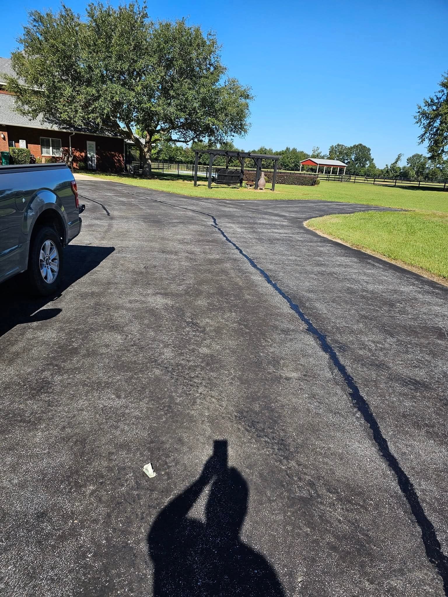 A paved driveway leads toward a tree and a small garden structure on a sunny day, with a vehicle parked on the left.