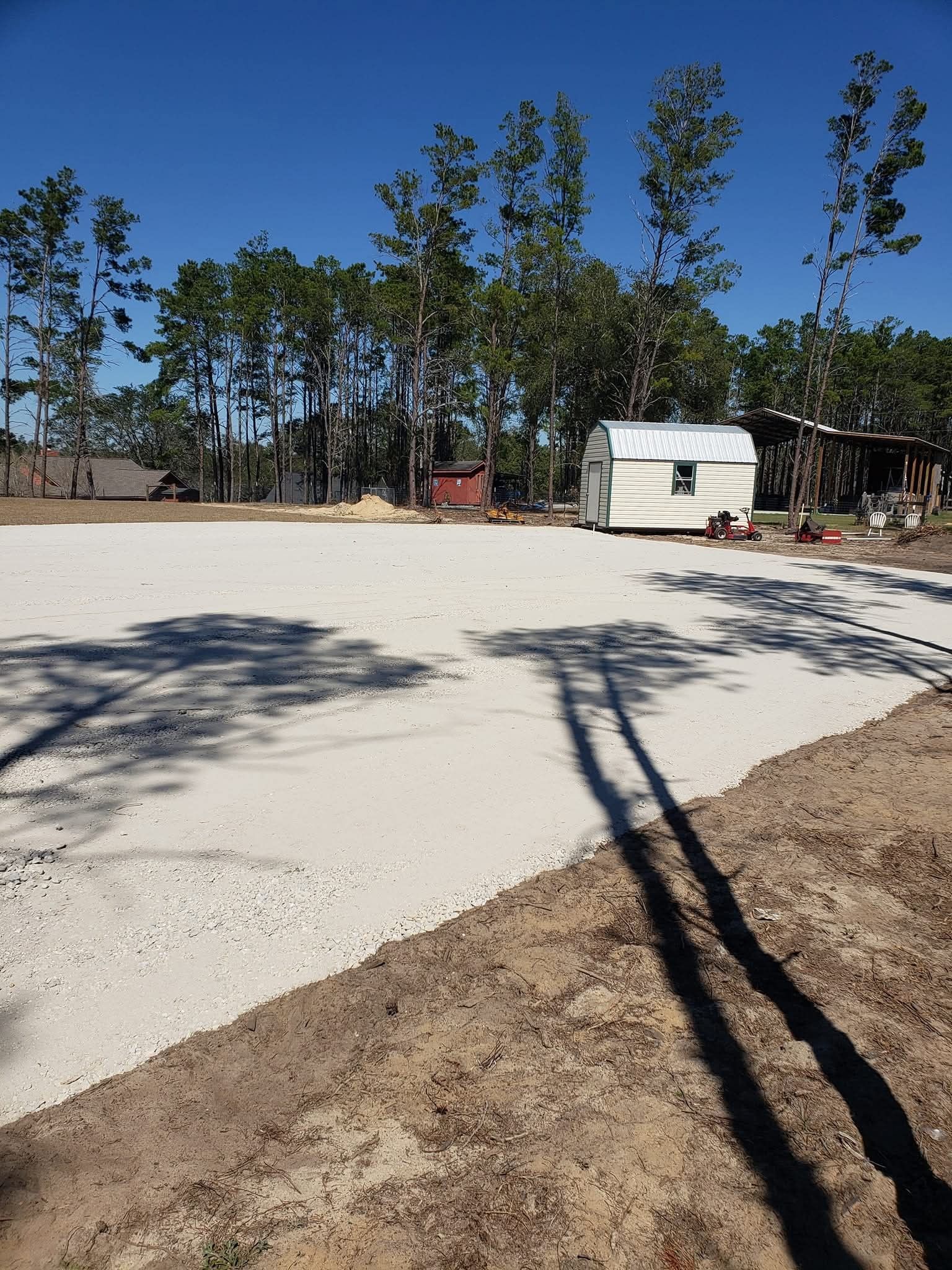 A wide, light-colored gravel pad sits in an open clearing, with a small shed and trees in the background under a blue sky.
