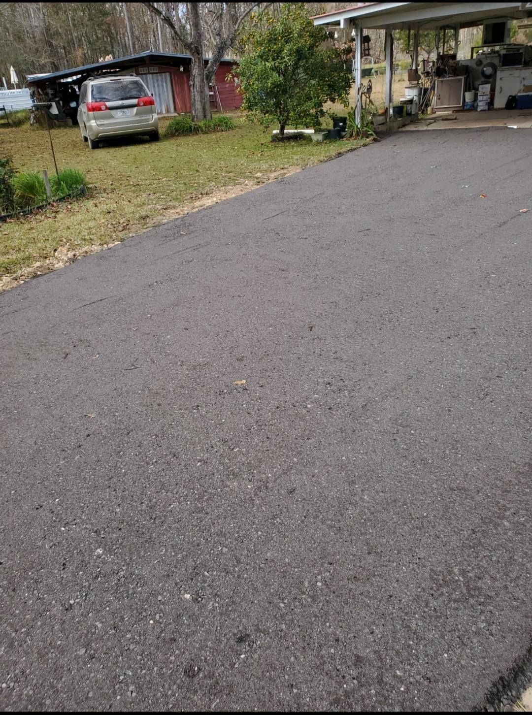 A paved driveway leads toward a house with a carport and a silver SUV parked in the yard on a cloudy day.