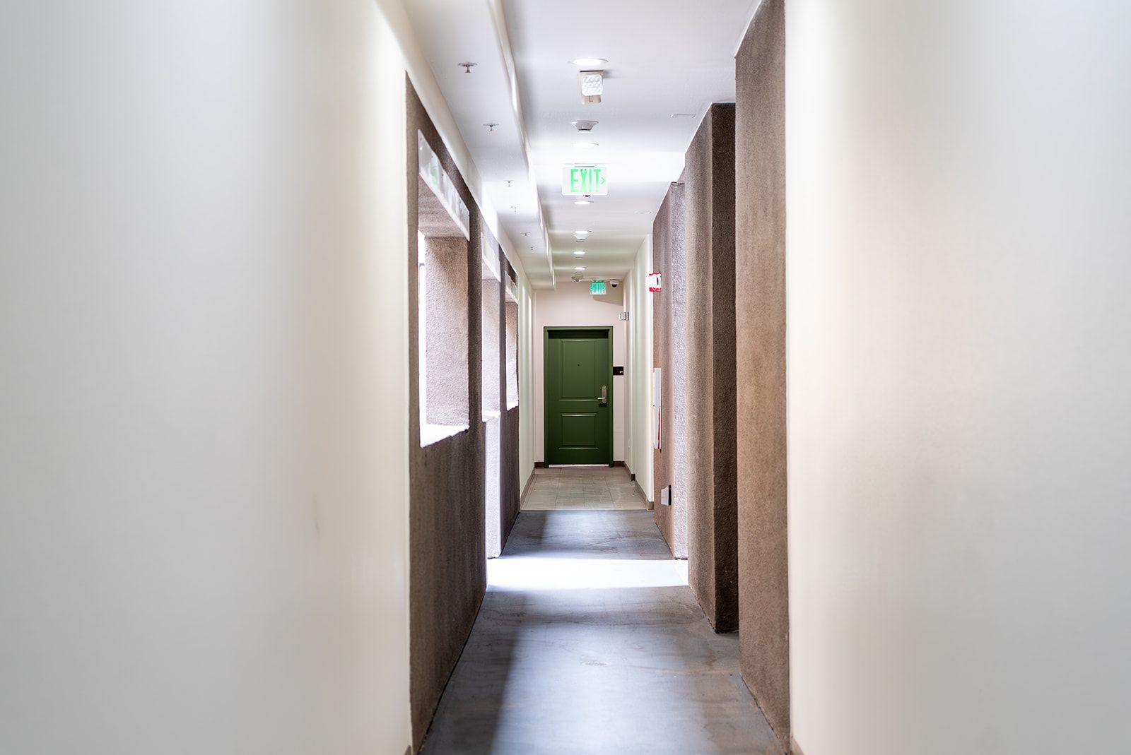 Narrow indoor hallway with white walls, windows, and a green door at the end.