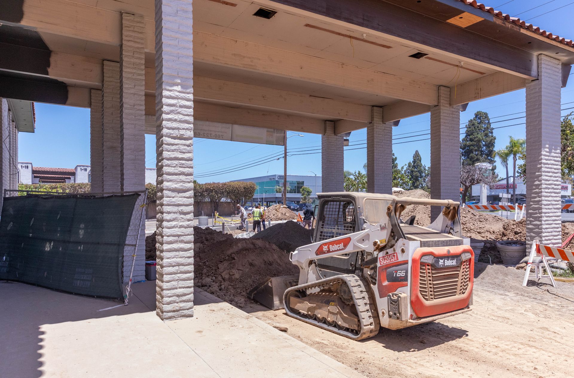 Construction site with a compact loader parked under a building overhang beside stone columns.