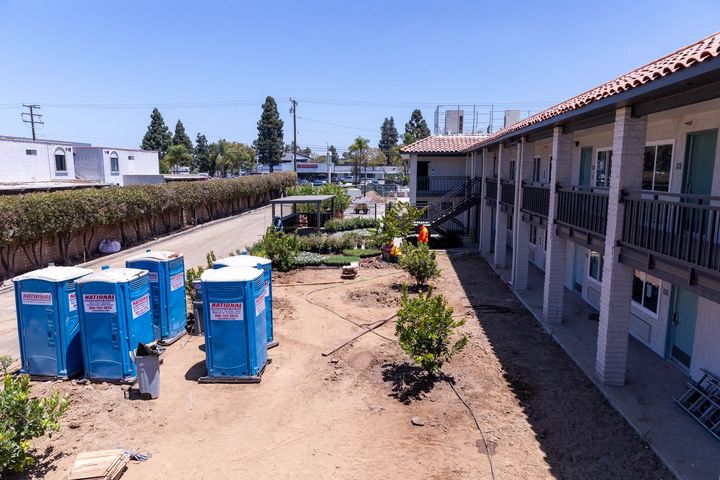Outdoor walkway beside a building with blue portable toilets and potted plants in bright sunlight