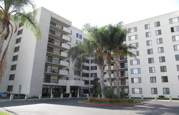 Apartment complex with white multi-story buildings, palm trees, and a landscaped courtyard.