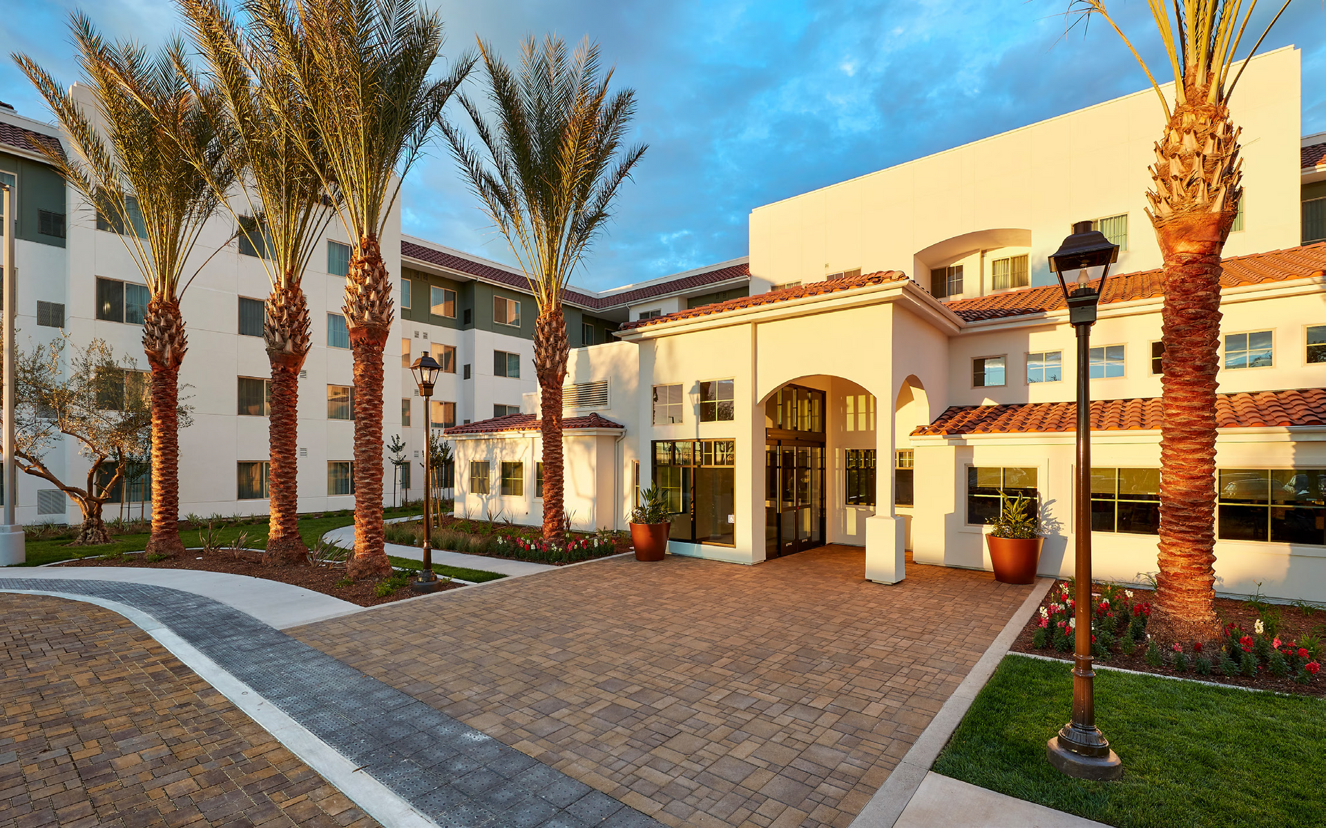 Modern apartment complex entrance with palm trees, white buildings, and a paved walkway at sunset