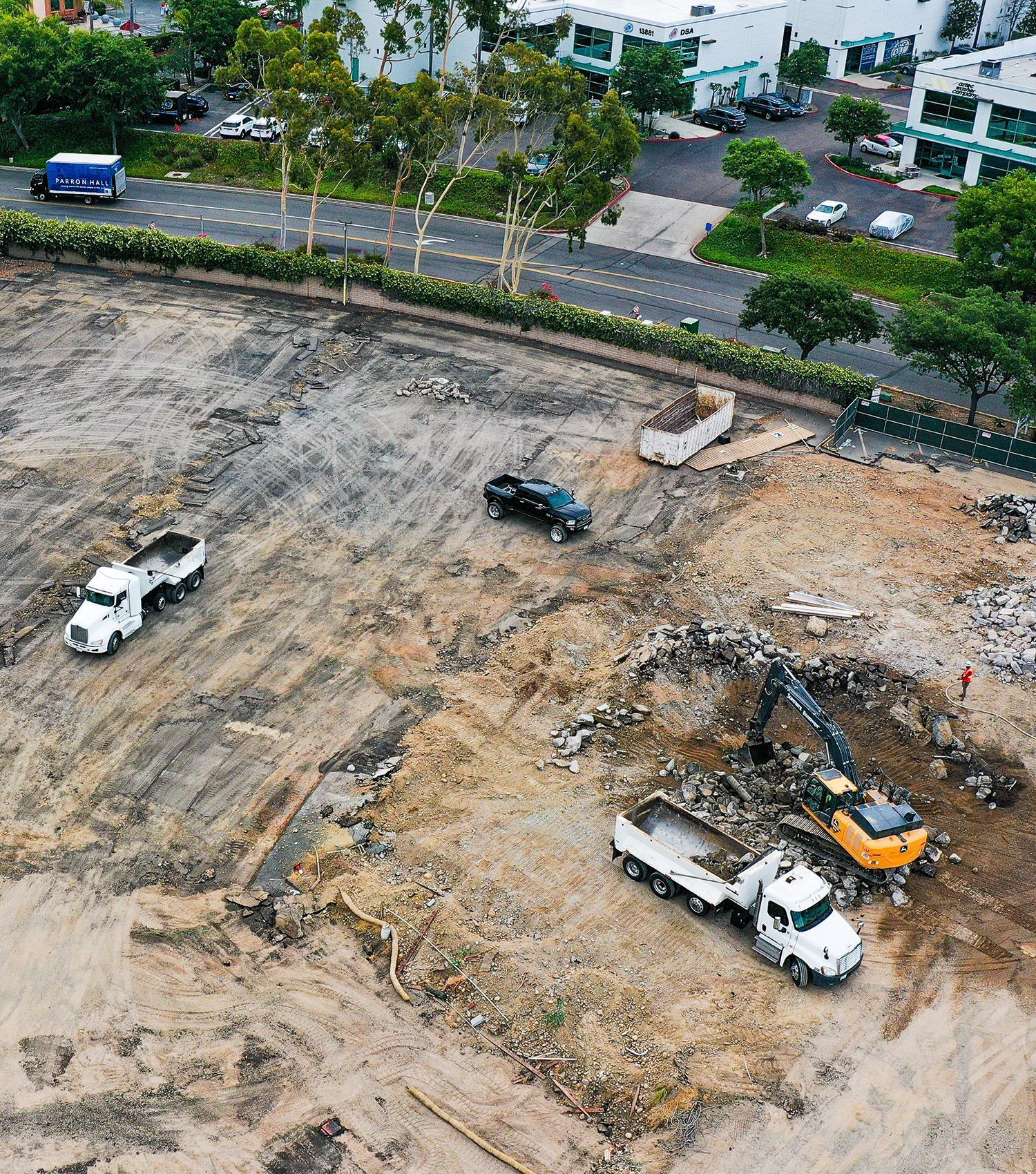Aerial view of a construction site with excavators, dump trucks, and bare earth beside a road and buildings