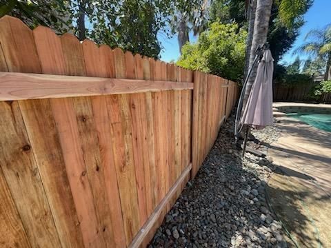 Wooden privacy fence along a poolside walkway, with rocks and a closed patio umbrella nearby.