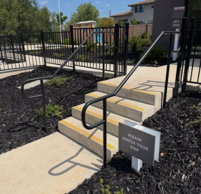 Concrete accessibility ramp and steps with black railings beside a fenced apartment walkway.