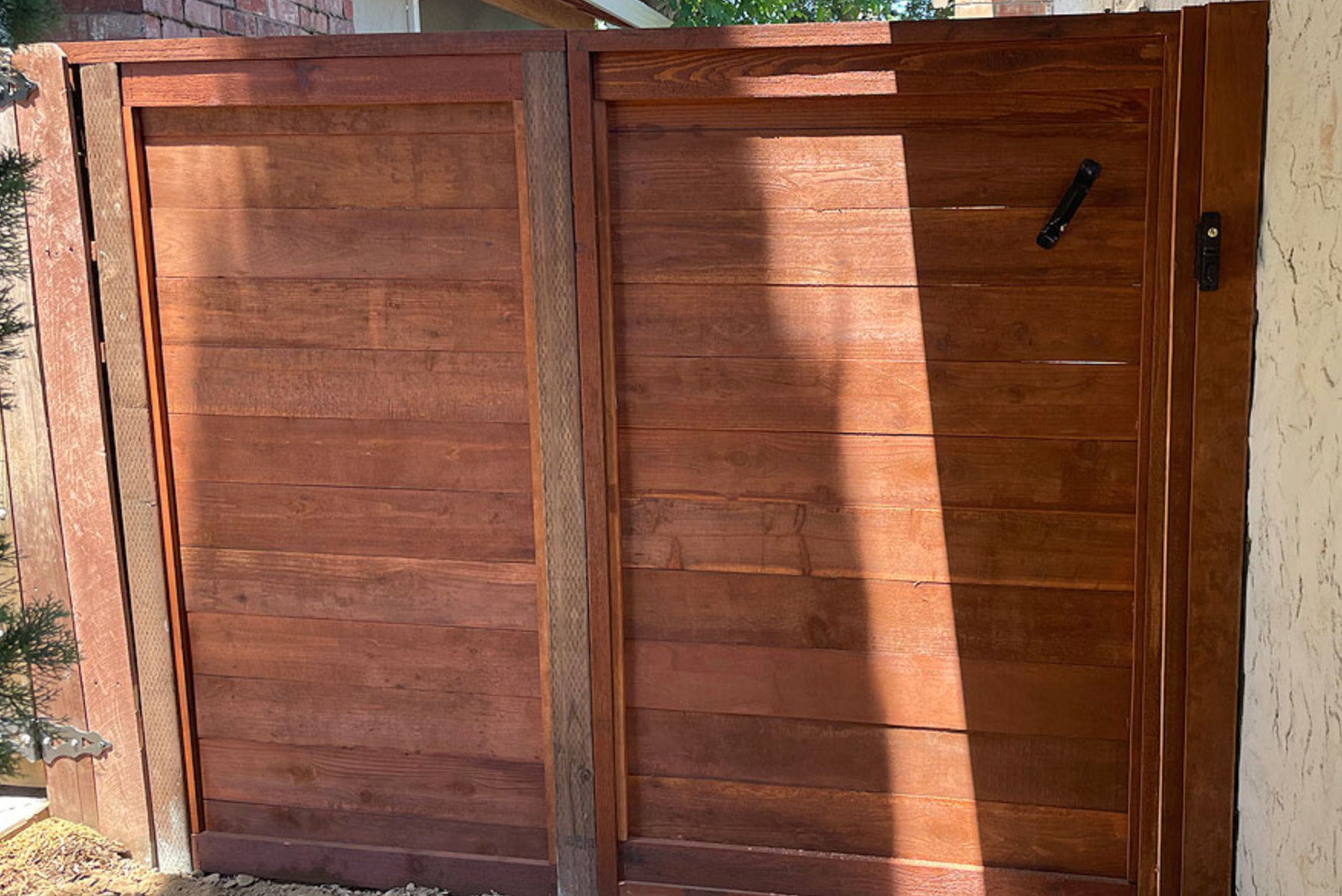 Wooden double gate, stained brown, with black hardware. Sunlight casts shadows on the planks.
