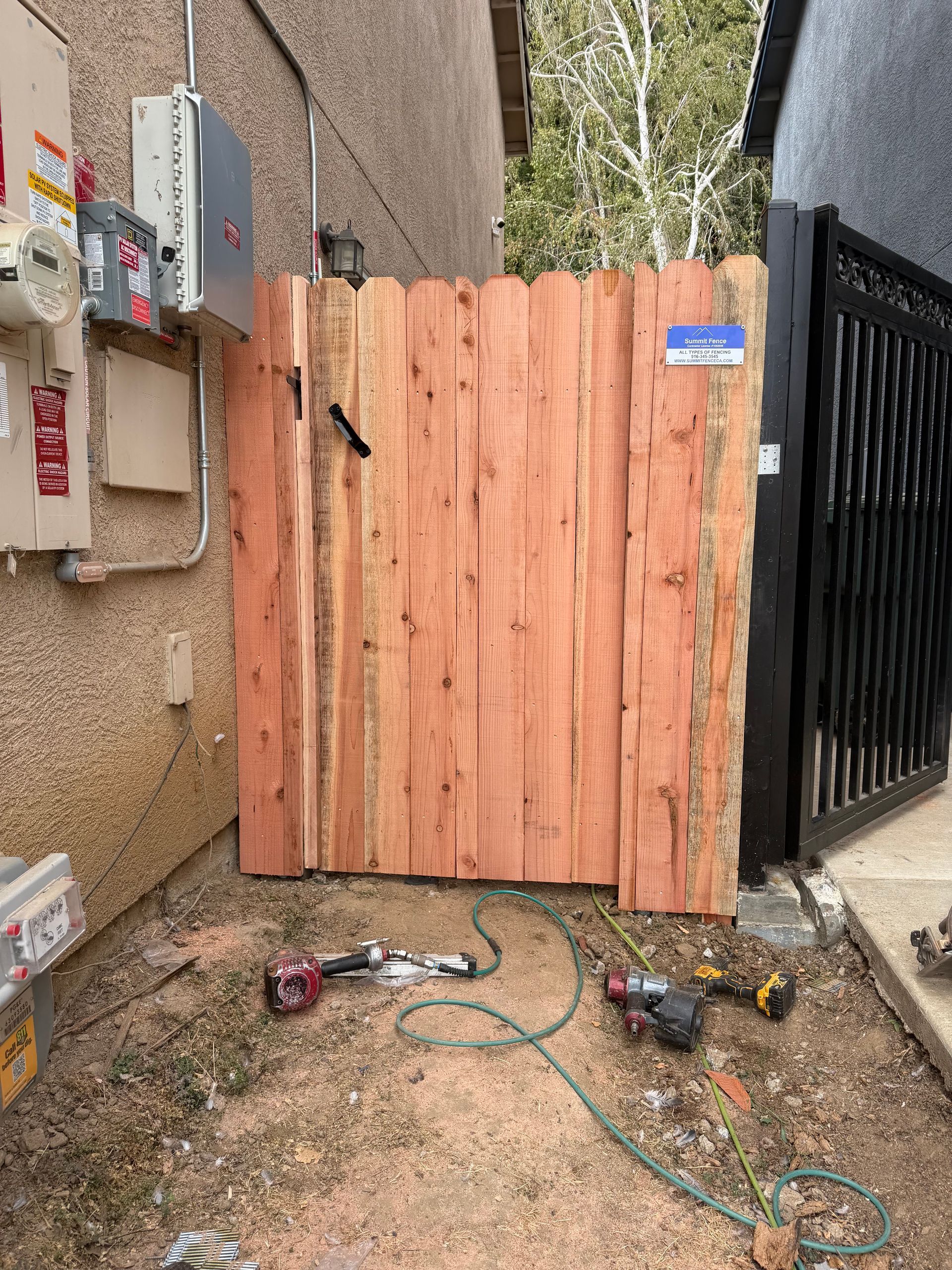 Wooden gate between buildings with tools and extension cords on gravel below