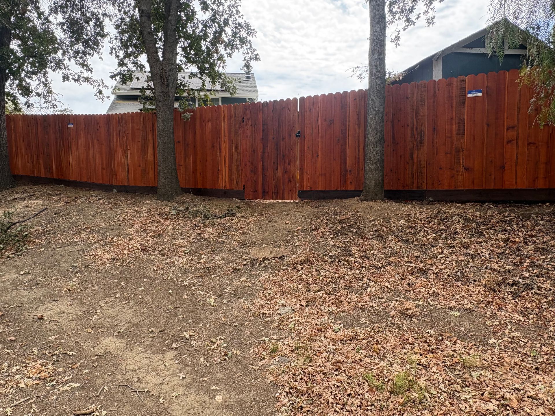 Red wooden fence along a leaf-covered yard with trees and houses behind it