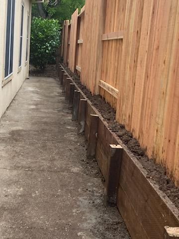 Narrow concrete walkway beside a house and tall wooden fence, with raised garden beds along the fence.